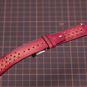Puede incluir: Una correa de reloj de cuero burdeos con un dise&ntilde;o perforado. La correa tiene costuras blancas a lo largo de los bordes y una hebilla plateada. La correa del reloj mide aproximadamente 23 cm de largo.