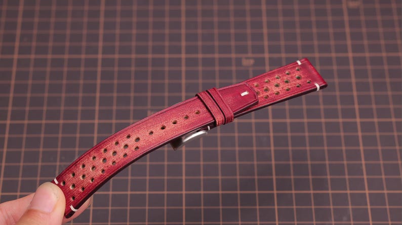 Puede incluir: Una correa de reloj de cuero burdeos con un dise&ntilde;o perforado. La correa tiene costuras blancas a lo largo de los bordes y una hebilla plateada. La correa del reloj mide aproximadamente 23 cm de largo.