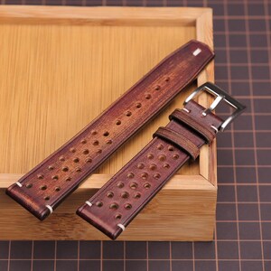 Puede incluir: Dos correas de reloj de cuero marr&oacute;n con un dise&ntilde;o perforado y costuras blancas. Una correa tiene una hebilla plateada. Las correas se exhiben en una bandeja de madera, mostrando su rico color y textura. Un accesorio elegante.