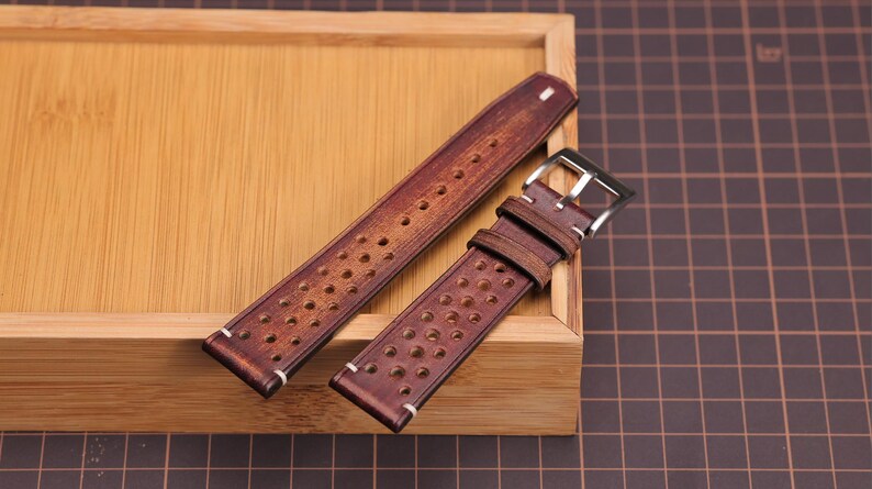 Puede incluir: Dos correas de reloj de cuero marr&oacute;n con un dise&ntilde;o perforado y costuras blancas. Una correa tiene una hebilla plateada. Las correas se exhiben en una bandeja de madera, mostrando su rico color y textura. Un accesorio elegante.