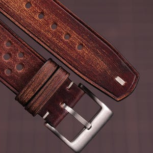 Puede incluir: Primer plano de una correa de reloj de cuero marr&oacute;n con una hebilla plateada. La correa tiene un acabado envejecido y presenta m&uacute;ltiples perforaciones circulares. Las costuras blancas acent&uacute;an los bordes y la hebilla es rectangular.
