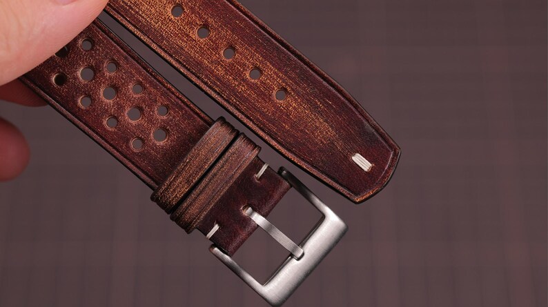 Puede incluir: Primer plano de una correa de reloj de cuero marr&oacute;n con una hebilla plateada. La correa tiene un acabado envejecido y presenta m&uacute;ltiples perforaciones circulares. Las costuras blancas acent&uacute;an los bordes y la hebilla es rectangular.
