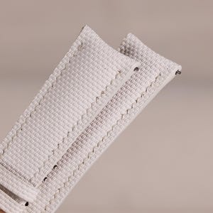 Puede incluir: Primer plano de una correa de reloj blanca con un patrón de tejido texturizado. La correa tiene forma rectangular con bordes cosidos y herrajes metálicos para la fijación. La correa está sujeta por una mano.