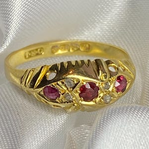 Puede incluir: Un anillo de oro con un diseño detallado que presenta tres rubíes rojos y diamantes blancos más pequeños. El anillo está marcado con "9ct" en la banda interior.