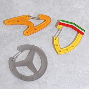 May include: Three carabiner clips are shown. One is orange, another is yellow with red, white, and green stripes, and the third is grey. Each clip has a metal spring-loaded gate. These are suitable for various outdoor and everyday uses.