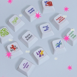 Puede incluir: Una colección de teclas blancas para teclado mecánico con varias frases y gráficos impresos, incluyendo "simply lovely", "forza leclerc", "it's hammer time", "Nothing just an inchident", "NORRIS", "LH 44", "AK12" y "mamma mia".