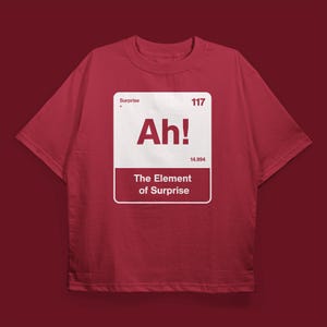 May include: A maroon t-shirt with a white graphic. The graphic resembles a periodic table element, with the word "Ah!" in large letters and the phrase "The Element of Surprise" below. The shirt has a crew neck.
