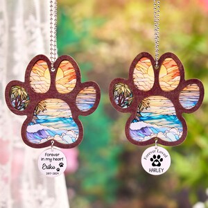 May include: Two paw-shaped ornaments with a beach scene design. The ornaments are made of wood and feature a colorful sunset, ocean waves, and palm trees. Each ornament has a small, round tag with a name and the phrase "Forever in my heart".