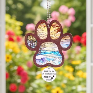 May include: A wooden paw print ornament with a beach scene design. The paw print is brown with a stained glass style image of a beach, ocean, and sunset. A small circular tag reads "Look For Me In The Rainbows Jasper 2016-2025".