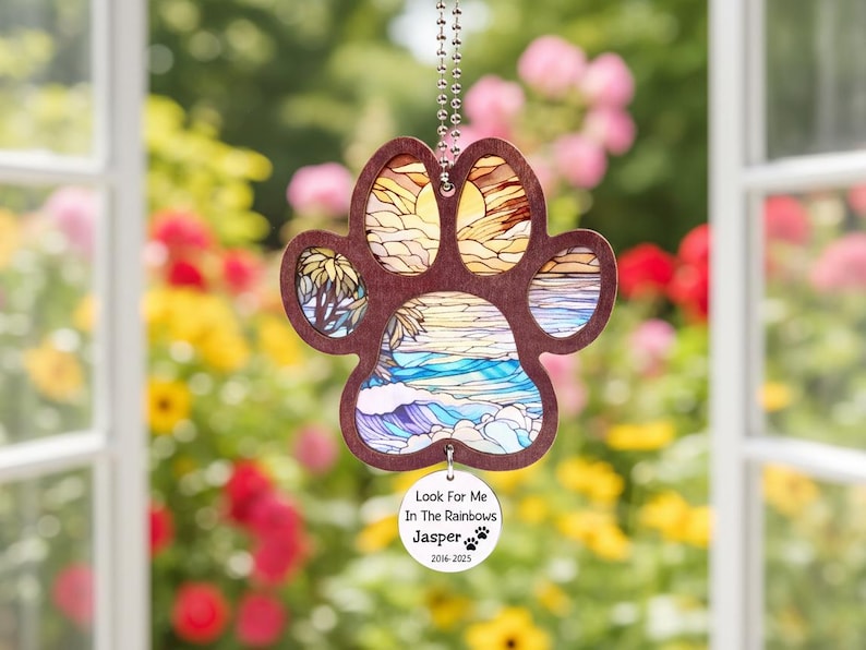 May include: A wooden paw print ornament with a beach scene design. The paw print is brown with a stained glass style image of a beach, ocean, and sunset. A small circular tag reads "Look For Me In The Rainbows Jasper 2016-2025".