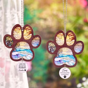 May include: Two paw-shaped car charms with a beach scene design. The charms have a brown border and a colorful interior depicting a sunset over the ocean. Each charm has a small, round tag with a pet's name and a quote. The text on one charm says "Look for me in the rainbows Simba".