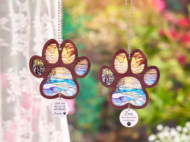 May include: Two paw-shaped car charms with a beach scene design. The charms have a brown border and a colorful interior depicting a sunset over the ocean. Each charm has a small, round tag with a pet's name and a quote. The text on one charm says "Look for me in the rainbows Simba".