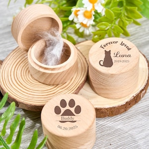 May include: Wooden pet memorial urns. One urn is open, revealing pet fur. Two other urns have engraved designs: a cat silhouette with "Forever Loved Luna 2019-2025" and a paw print with "Buddy 06.20.2025".