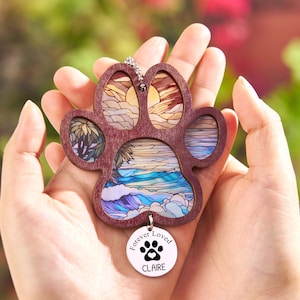 May include: A wooden paw-shaped pendant with a stained-glass design of a beach scene. The pendant has a small, round charm that reads "Forever Loved" with a paw print and the name "Claire".