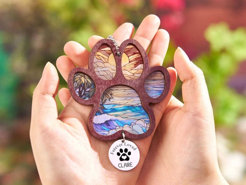 May include: A wooden paw-shaped pendant with a stained-glass design of a beach scene. The pendant has a small, round charm that reads "Forever Loved" with a paw print and the name "Claire".