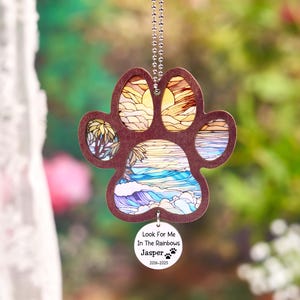 May include: A wooden paw print ornament with a beach scene design. The paw print is brown with a stained glass effect showing a sunset, palm trees, and ocean waves. A small, round tag reads "Look For Me In The Rainbows" and "Jasper 2016-2025".