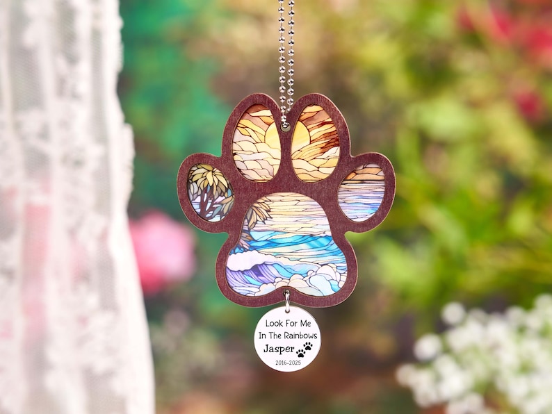 May include: A wooden paw print ornament with a beach scene design. The paw print is brown with a stained glass effect showing a sunset, palm trees, and ocean waves. A small, round tag reads "Look For Me In The Rainbows" and "Jasper 2016-2025".