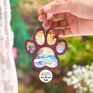 May include: A wooden paw print ornament with a beach scene design. The ornament has a brown border and a chain for hanging. The beach scene includes a sunset, palm trees, and ocean waves. A small white tag reads "IN LOVING MEMORY OF" and the name "Bella".