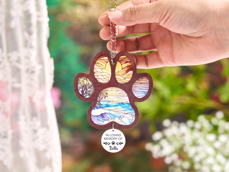 May include: A wooden paw print ornament with a beach scene design. The ornament has a brown border and a chain for hanging. The beach scene includes a sunset, palm trees, and ocean waves. A small white tag reads "IN LOVING MEMORY OF" and the name "Bella".