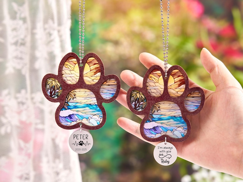 May include: Two wooden paw print ornaments with a beach scene design. Each ornament has a silver chain and a circular tag with text. One tag says "Peter" and the other says "I'm always with you Ruby".