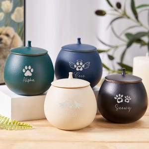 Op de afbeelding: Vier keramische urnen voor huisdieren met gepersonaliseerde gravures. De urnen zijn in groen, blauw, crème en zwart. De gravures omvatten poot afdrukken, engelenvleugels en namen: Alpha, Coco, Snowy.