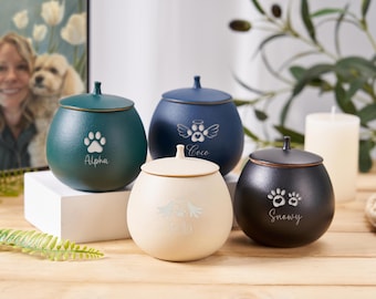 May include: Four ceramic pet urns with personalized engravings. The urns are in green, blue, cream, and black. The engravings include paw prints, angel wings, and names: Alpha, Coco, Snowy.
