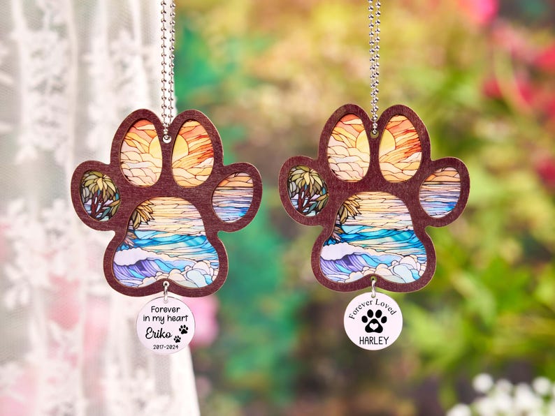 May include: Two paw-shaped ornaments with a beach scene design. The ornaments are made of wood and feature a colorful sunset, ocean waves, and palm trees. Each ornament has a small, round tag with a name and the phrase "Forever in my heart".