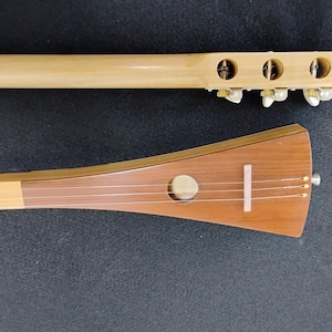 May include: A close-up of a traditional Japanese shamisen. The instrument features a wooden body with a brown soundboard, a long neck, and three strings. The headstock has tuning pegs. The shamisen is a plucked string instrument.