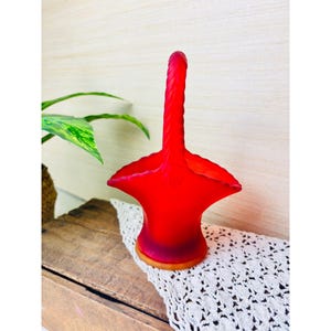 Peut inclure: Un panier en verre dépoli rouge vif doté d'une anse haute torsadée et d'un large bord festonné. La base du panier présente une teinte rouge orangé légèrement plus claire, avec une texture mate et lisse sur l'ensemble de l'objet.
