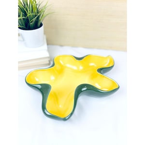 Könnte beinhalten: Eine gelb-grüne Keramikschale in einer einzigartigen, abstrakten Form. Das Innere ist leuchtend gelb, während der Rand und die Außenseite dunkelgrün sind. Die Schale dient wahrscheinlich zum Servieren oder zur Präsentation.