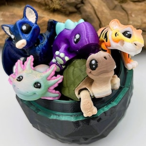 May include: A selection of vibrant 3D-printed animal figures, including a blue bat, purple dinosaur, orange tiger, green turtle, and pink axolotl, presented in a black container with green edging. The figures feature large, cartoonish eyes.