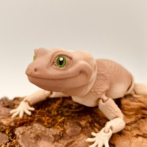 Könnte beinhalten: Eine hellbraune, bewegliche Echsenfigur mit strukturierter Oberfläche und leuchtend grünen Augen. Die Echse hat einen glatten, abgerundeten Kopf und sichtbare Gelenke an den Gliedmaßen. Sie ist auf einem Stück rauer, brauner Baumrinde platziert.