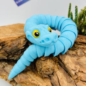 Könnte beinhalten: Ein hellblaues, bewegliches Schlangenspielzeug mit gelben Augen, das auf einem Holzstück platziert ist. Der Schlangenkörper ist für flexible Bewegungen segmentiert. Der Kopf hat ein detailliertes Design mit sichtbaren Nasenlöchern und einem lächelnden Mund.