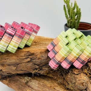 Könnte beinhalten: Zwei Stapel bunter, gestreifter Untersetzer liegen auf einem Holzstück. Ein Stapel hat rosa, rote und grüne Streifen, während der andere grüne, rosa und orangefarbene Streifen hat. Im Hintergrund befindet sich ein kleiner Kaktus in einem Topf.