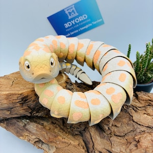 Könnte beinhalten: Ein 3D-gedrucktes, bewegliches Schlangenspielzeug mit beigefarbener Grundierung und orangefarbenen Flecken. Die Schlange hat einen detaillierten Kopf mit großen Augen und einen segmentierten Körper, der sich kreisförmig biegt. Eine blaue Karte mit dem Text 3DYORD ist im Hintergrund zu sehen.