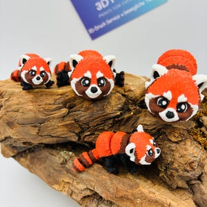 Könnte beinhalten: Eine Sammlung von 3D-gedruckten Rotpanda-Figuren in verschiedenen Posen. Die Figuren haben orangefarbene und braune Körper mit weißen Akzenten auf Gesicht und Ohren. Sie werden auf einem Holzstück präsentiert.