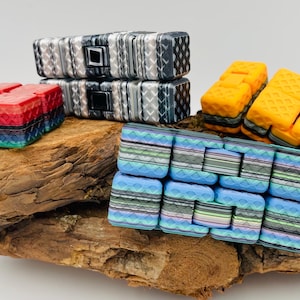 Könnte beinhalten: Vier bunte Fidget-Spielzeuge aus ineinandergreifenden Quadraten. Die Spielzeuge sind in verschiedenen Farben und Mustern gehalten, darunter Rot, Blau, Gelb und Schwarz. Die Spielzeuge sind auf einem Stück Treibholz angeordnet.