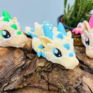 Könnte beinhalten: Drei skurrile Drachenfiguren in Beige, mit grünen, blauen und rosa Akzenten. Die Drachen sind auf einem Holzstück platziert, mit einer Pflanze im Hintergrund. Jeder Drache hat große, ausdrucksstarke Augen.