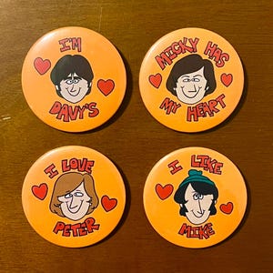 Könnte beinhalten: Vier orange runde Buttons mit Cartoon-Illustrationen von Gesichtern und Text. Jeder Button hat ein anderes Gesicht und einen anderen Text: "I'm Davy's", "Micky Has My Heart", "I Love Peter" und "I Like Mike". Jeder Button hat auch rote Herzgrafiken.