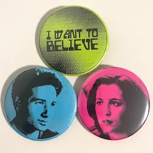 Può includere: Tre spille rotonde. Una è verde lime con il testo "I WANT TO BELIEVE" in nero. Le altre due spille presentano ritratti, uno in blu e nero, l'altro in rosa e nero.