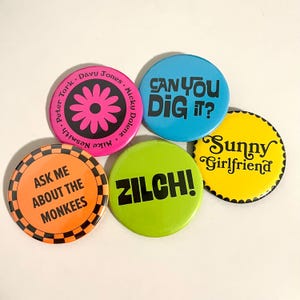 Puede incluir: Un conjunto de cinco botones redondos y coloridos. Un botón rosa con un diseño floral y los nombres de The Monkees. Un botón azul dice "CAN YOU DIG IT?" y un botón amarillo dice "Sunny Girlfriend". Un botón naranja dice "ASK ME ABOUT THE MONKEES" y un botón verde dice "ZILCH!"