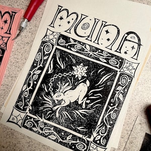May include: A black and white print featuring the word "Muna" in stylized lettering. The print depicts a rabbit with antlers and a flail within a decorative frame of vines and geometric shapes. A red-handled carving tool is visible.