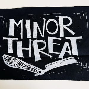 Peut inclure: Écusson en tissu noir avec les mots "MINOR THREAT" en lettres blanches et grasses. Une illustration blanche d'un rasoir droit est en dessous du texte. L'écusson est de style linogravure, imprimé à la main.
