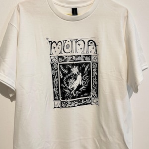 May include: A white t-shirt with a black graphic print. The print features the word "MUNA" in a decorative font above a framed illustration of a figure with floral and vine elements. The shirt is hanging on a gold hanger.