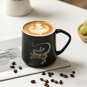Puede incluir: Una taza de café de cerámica negra con borde blanco, llena de un café con leche con un diseño decorativo de espuma. La taza tiene un gráfico de esqueleto de dinosaurio dorado. Los granos de café están esparcidos alrededor de la taza.