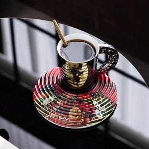 以下が含まれることがあります： 黒い陶器製のコーヒーカップとソーサーのセット。反射するアナモルフィックデザインが施されており、側面から見るとカボチャ、骸骨、幽霊などのカラフルなハロウィーンのイラストが浮かび上がります。金色のスプーンがカップの中に入っています。