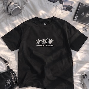May include: A black short-sleeved t-shirt featuring a white graphic of three stylized star shapes centered on the chest. Below the stars, the text +OMORROW X +OGETHER is printed in a clean, white sans-serif font. The shirt has a crew neckline.