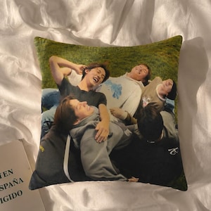 May include: A square throw pillow featuring a printed photograph of four individuals lying on a grassy field. The image shows the group in casual clothing, including hoodies and t-shirts, captured from an overhead perspective while resting outdoors.