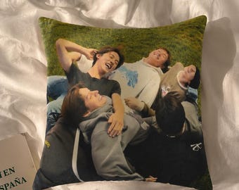 Puede incluir: Un cojín cuadrado con una fotografía impresa de cuatro personas tumbadas en un campo de hierba. La imagen muestra al grupo con ropa informal, incluyendo sudaderas con capucha y camisetas, captado desde una perspectiva cenital.