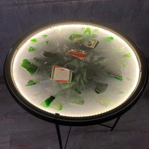 Pode incluir: Uma mesa redonda de vidro com uma moldura de metal preto e três pernas. A superfície da mesa está cheia de pedaços de garrafas de vidro verde quebradas e tem um acabamento de resina branca e nublada. A superfície da mesa é iluminada com um anel de luzes LED brancas ao redor da borda. A superfície da mesa também tem dois pequenos rótulos retangulares vermelhos e brancos que dizem "Jägermeister" e "56".